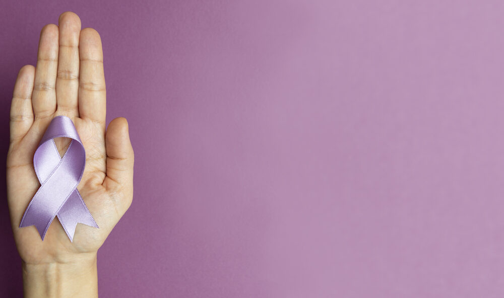 A hand holds a lilac silk ribbon in a loop against a purple background, representing Alzheimer's Awareness logo.